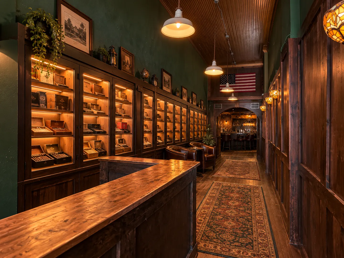 Interior of a traditional cigar lounge with a dark-stained wooden counter, glass-enclosed humidor cabinets with warm lighting, forest-green wainscoted walls, leather armchairs, and an arched opening to a brick bar area