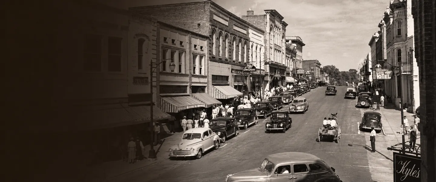 Sepia-toned vintage photograph of a busy Main Street with parked cars, pedestrians, and historic brick storefronts